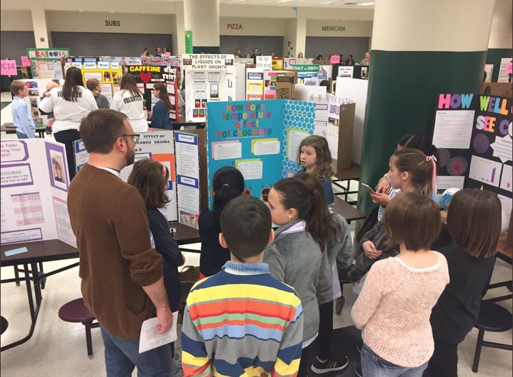 A group of science fair attendees admire and attentively listen to s participant’s project on the effect of temperature on the taste of hot chocolate.
